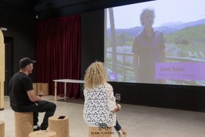 Dos visitants del Centre Enoturístic Pla de Bages asseguts visualitzant un vídeo mentre prenen una copa de vi