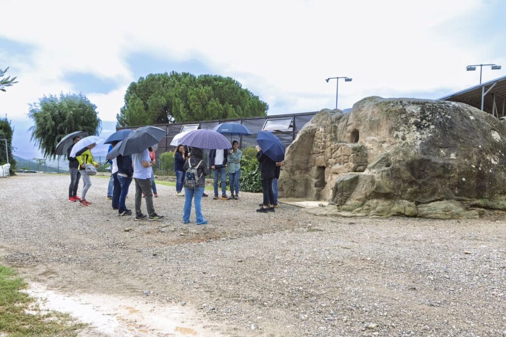 Grup de visitants amb paraigües davant la tina medieval de pedra al Celler Can Serra dels Exibis, DO Pla de Bages