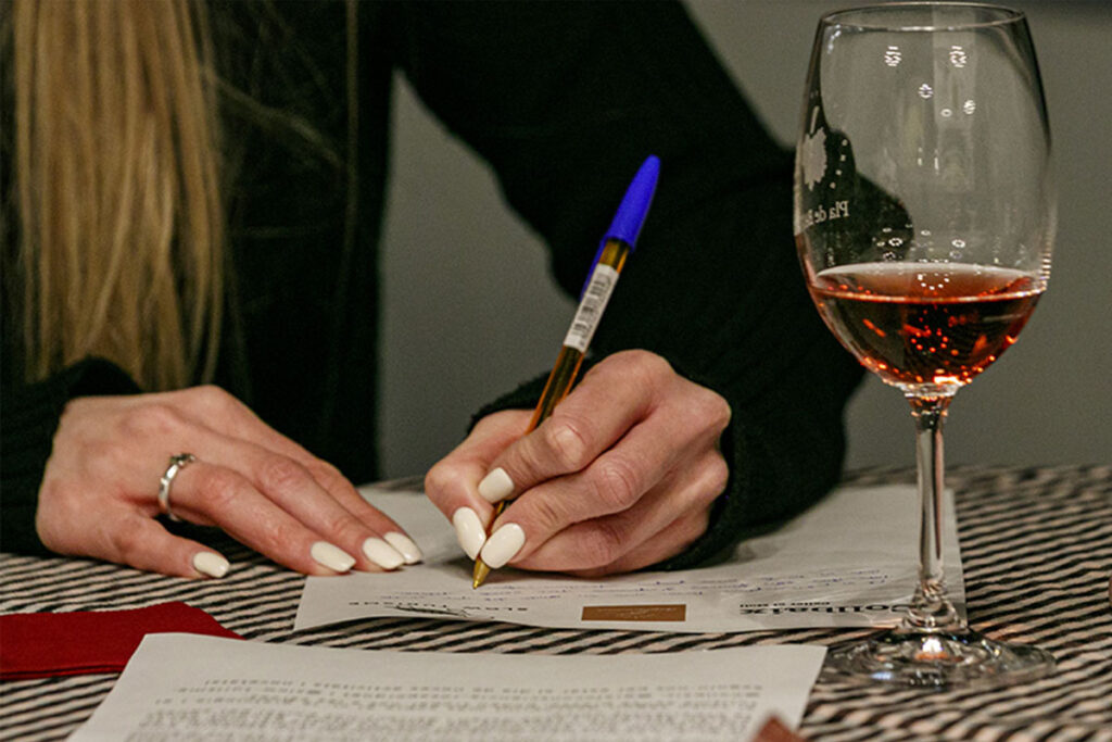 Mà d’una dona escrivint en un full durant un tast de grafologia amb copa de vi rosat del Collbaix Celler el Molí, DO Pla de Bages.