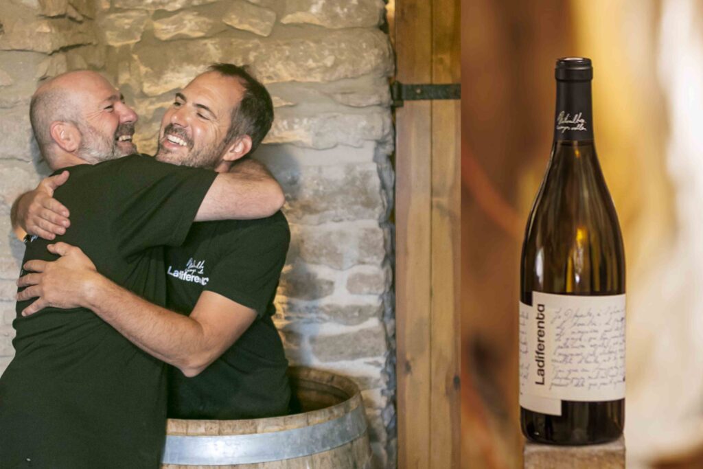 Joan i Martí de Ladiferenta abraçant-se dins el celler amb una ampolla del seu vi blanc, projecte de la DO Pla de Bages